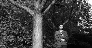 A black and white photograph of a woman standing beside a tall tree